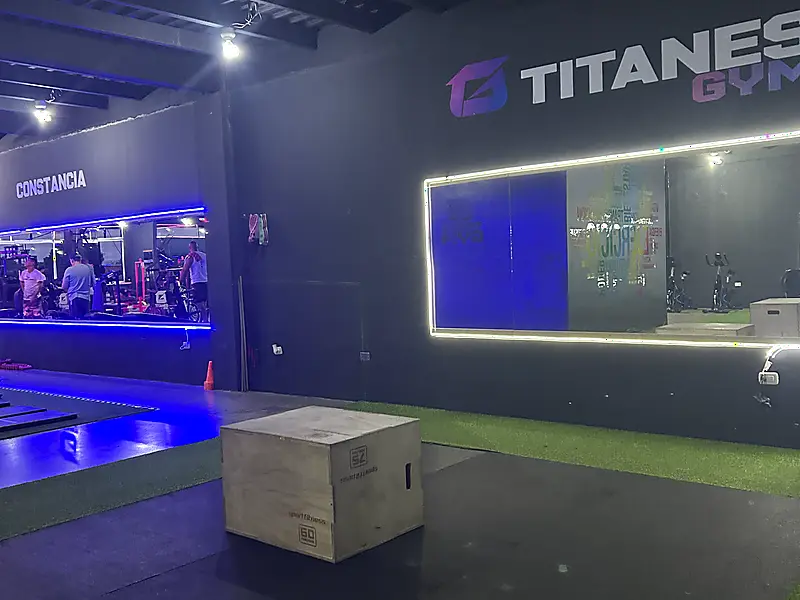 Pared iluminada con el logo de Titanes Gym junto al área de pesas libres